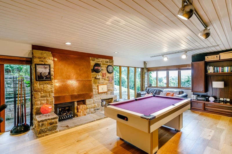Dual-aspect games room with oak flooring, partial floor to ceiling glazing, French doors out to the side aspect, fitted console units with shelving and storage and a focal point, stone-built fireplace with a copper canopy and inset gas fire.