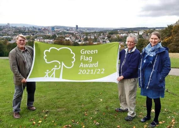 Councillor Alison Teal stands with Cactus from Meersbrook Park Users Trust, proudly holding the Green Flag award over the views of Meersbrook Park with Councillor Brian Holmshaw.