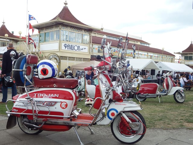 The local International Scooter festival in Ryde on the IOW back on 2014. Scooters parked outside the Pavilion, 3 Mods admire a scooter and have a drink and a chat.