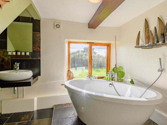 A beautifully-appointed family bathroom that boasts a contemporary four-piece suite, the highlight of which is a large, oval-shaped bath-tub. There is a also a separate, tiled shower-enclosure with a 'rain' shower and an oval-shaped sink mounted on a wall-hung work surface. A slate tiled floor and beamed ceiling add to the room's appeal.