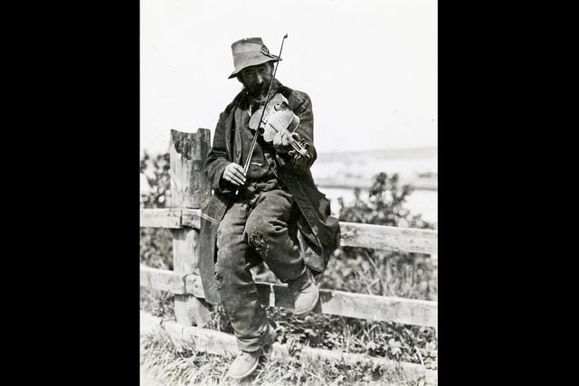 A travelling fiddler plays a tune on a fence. The photographer, originally from Galloway, was an electrical engineer who became interested in social welfare while working in London, where he volunteered at a Cockney Mission.