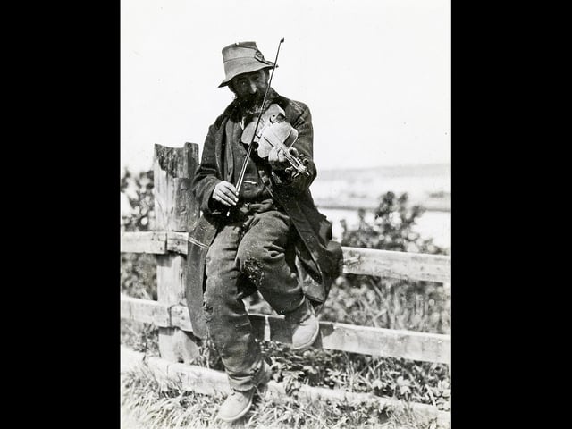 A travelling fiddler plays a tune on a fence. The photographer, originally from Galloway, was an electrical engineer who became interested in social welfare while working in London, where he volunteered at a Cockney Mission.