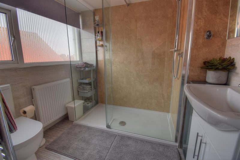 With a side-facing window, tiled walls, tiled floor, large walk-in shower unit, low-flush WC, hand basin with vanity unit, heated towel radiator and extractor fan.