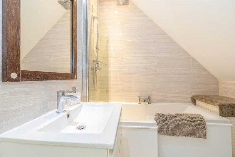 The contemporary-styled, luxury bathroom features an L-shaped bath, low flush WC, chrome heated towel rail and a square wash hand basin with storage beneath. Fitted over the bath is a chrome shower with a square, oversized, waterfall shower head.