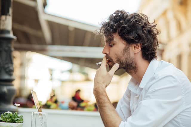 handsome elegant man sitting outside at a restaurant table, working and using laptop while smoking cigarettes