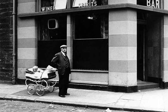 A man in cap stands beside a child in a pram outside a bar in Tollcross.