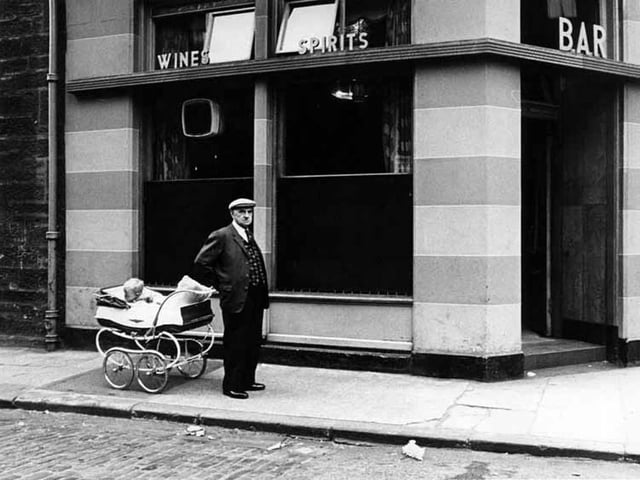 A man in cap stands beside a child in a pram outside a bar in Tollcross.
