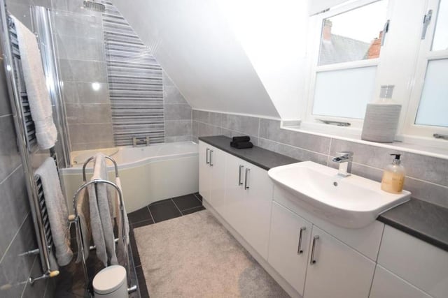 This contemporary en suite serves the main bedroom. It is fitted with a P-shaped, panelled bath that has a rainfall shower above, a low-flush WC and a wash basin in a vanity unit. The floor and walls are tiled, while there is also a chrome, heated towel-rail and warm, atmospheric downlighting.
