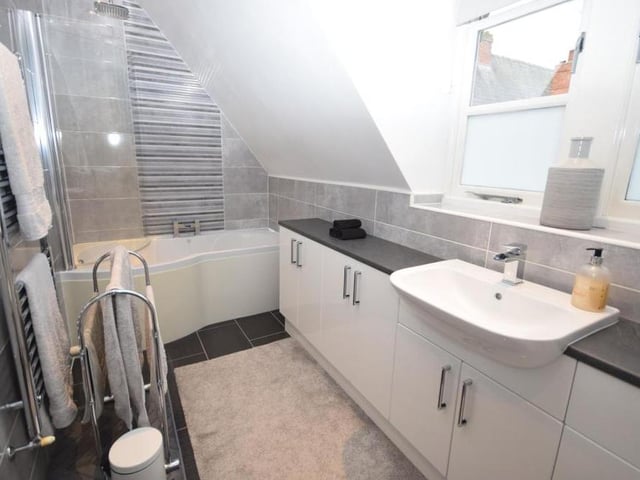 This contemporary en suite serves the main bedroom. It is fitted with a P-shaped, panelled bath that has a rainfall shower above, a low-flush WC and a wash basin in a vanity unit. The floor and walls are tiled, while there is also a chrome, heated towel-rail and warm, atmospheric downlighting.