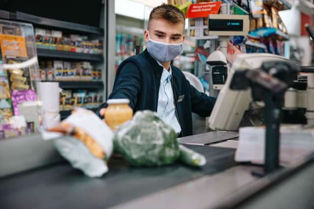 Young sales clerk wearing face mask sitting by cash register in supermarket and serving shoppers. Male cashier scanning grocery products at checkout.